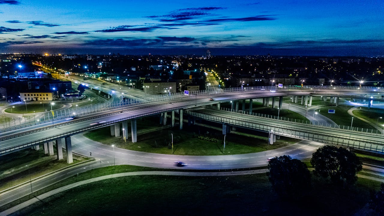 A dramatic cityscape featuring a modern flyover lit by vibrant city lights at night.